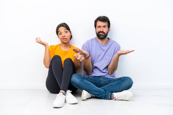 Young caucasian couple sitting on the floor isolated on white background having doubts and with confuse face expression - Valokuva, kuva