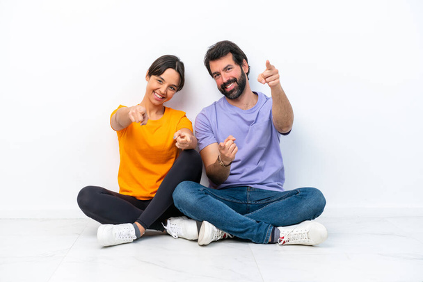 Young caucasian couple sitting on the floor isolated on white background points finger at you with a confident expression - Foto, afbeelding