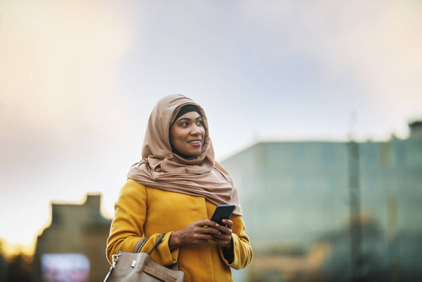 Zwarte moslim vrouw dragen hijab met een gelukkig gezicht staan in stedelijke omgeving en met behulp van haar smartphone. - Foto, afbeelding