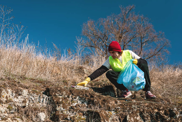 man nature activist crouches and stretches his hand towards empty glass bottle with blue trash bag - Zdjęcie, obraz