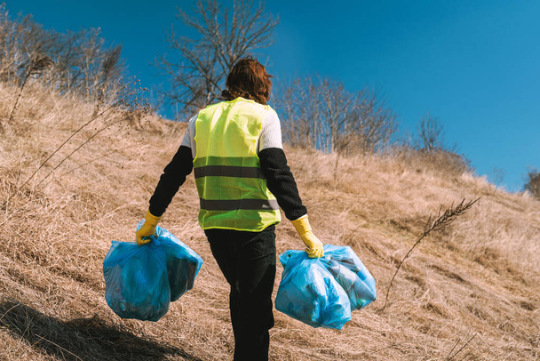 Sarı yelekli doğa eylemcisi güneşli tarla fotoğrafında mavi çöp torbalarını yol boyunca taşıyor. - Fotoğraf, Görsel