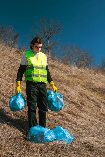 Uomo attivista della natura in giubbotto giallo tiene borse blu spazzatura lungo il percorso nel campo soleggiato - Foto, immagini