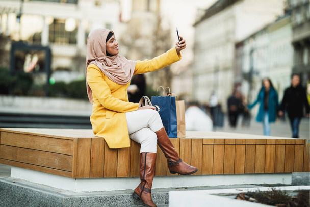 陽気黒いイスラム教徒の女性がヒジャーブを着て、彼女のスマートフォンで都会の環境で自撮り. - 写真・画像