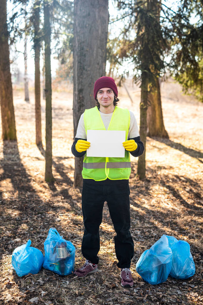 Mannelijke natuur activist houdt wit sjabloon papier terwijl staan in de buurt van blauwe vuilniszakken - Foto, afbeelding