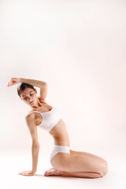 Studio shot of fit woman wearing seamless underwear sitting on the floor isolated on white. Slim female with her hair tied in ponytail posing in white lingerie. Copy space, close up, background. - Fotó, kép