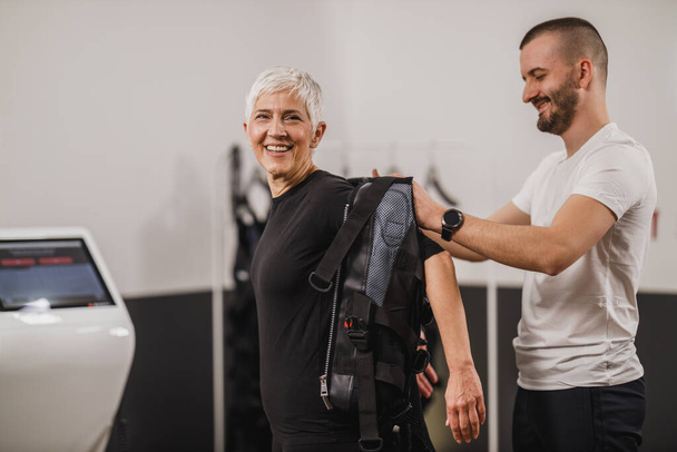 Personal Trainer hilft einer Seniorin im Anzug beim EMS-Training im Fitnessstudio. - Foto, Bild