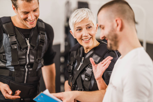 Sağlık görevlileri spor salonunda antrenman yapmadan önce orta yaşlı bir çift kişisel antrenörle konuşuyor.. - Fotoğraf, Görsel