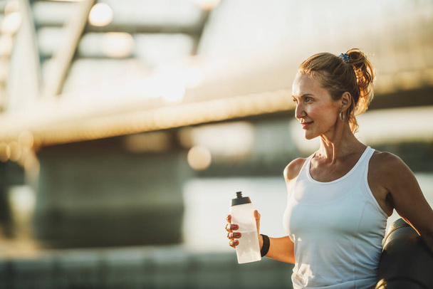 Middle age sportswoman holding water bottle before hard training near to river, in the city. - Foto, imagen