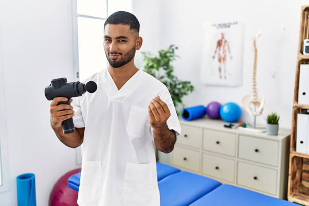 Young indian physiotherapist holding therapy massage gun at wellness center doing money gesture with hands, asking for salary payment, millionaire business  - Photo, Image