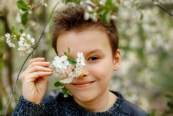 Happiness and excited kid enjoying spring. Adorable boy in spring blooming garden. International childrens day. The child is hidding his eye behind a flowering branch. The boy breathes easily. - Photo, Image