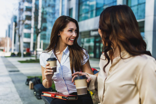 オフィスの外でコーヒーブレイク中におしゃべりする2人の笑顔のビジネス女性. - 写真・画像