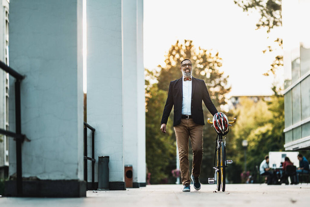 Επιτυχημένος μεσήλικας επιχειρηματίας πηγαίνει στη δουλειά με το ποδήλατο δίπλα του, περπατά μπροστά από το γραφείο περιοχή. - Φωτογραφία, εικόνα