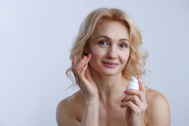 Portrait of adult woman with perfectly clean face skin applying moisturizing cream. Mid aged female with wavy blonde hair smiling performing daily skincare routine. Close up, copy space background. - Photo, Image