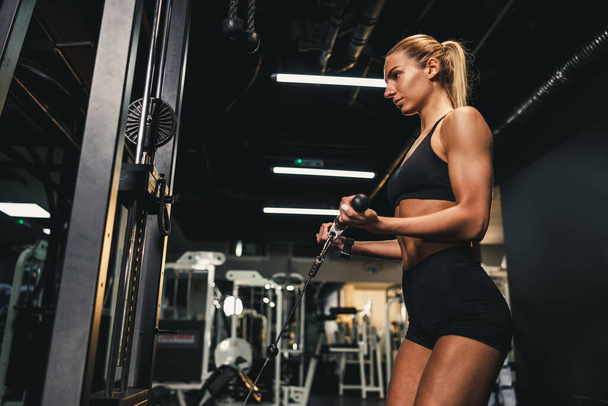 A young muscular woman is doing training on a machine in the gym. She is pumping up her biceps muscle with heavy weight. - Photo, Image