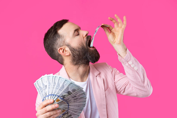 Portrait of a contented young businessman with a beard dressed in a pink jacket showing us dollar banknotes against a red studio background. Taste, smell of money. - Photo, Image