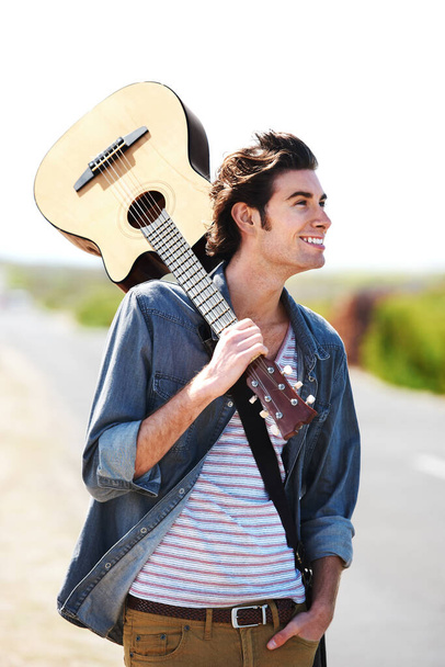 Es un verdadero muso. Sonriente joven sosteniendo su guitarra sobre su hombro de pie a un lado de la carretera - primer plano. - Foto, Imagen