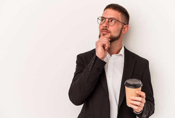 Junger kaukasischer Geschäftsmann hält Kaffee zum Mitnehmen isoliert auf weißem Hintergrund und blickt mit zweifelndem und skeptischem Gesichtsausdruck zur Seite. - Foto, Bild