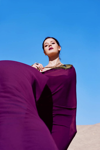 Niesamowita piękna brunetka z pawim piórem w fioletowej tkaninie na pustyni. Orientalny, indyjski, moda, koncepcja stylu - Zdjęcie, obraz