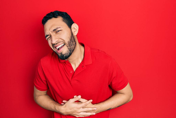 Hispanic man with beard wearing casual red t shirt with hand on stomach because nausea, painful disease feeling unwell. ache concept.  - Photo, image
