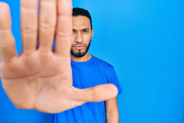 Spaanse man met baard draagt casual blauw t-shirt doen stoppen met zingen met palm van de hand. waarschuwingsuitdrukking met negatief en ernstig gebaar op het gezicht.  - Foto, afbeelding