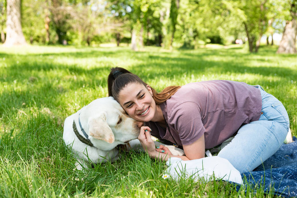 Νεαρή όμορφη γυναίκα στο πάρκο με το κατοικίδιο σκυλί της λευκό Λαμπραντόρ Retriever που υιοθετεί από το καταφύγιο ζώων για να του δώσει καλή ζωή και αγάπη. Γυναίκα λάτρης των ζώων με υιοθετημένο κουτάβι της διασκεδάζουν. - Φωτογραφία, εικόνα