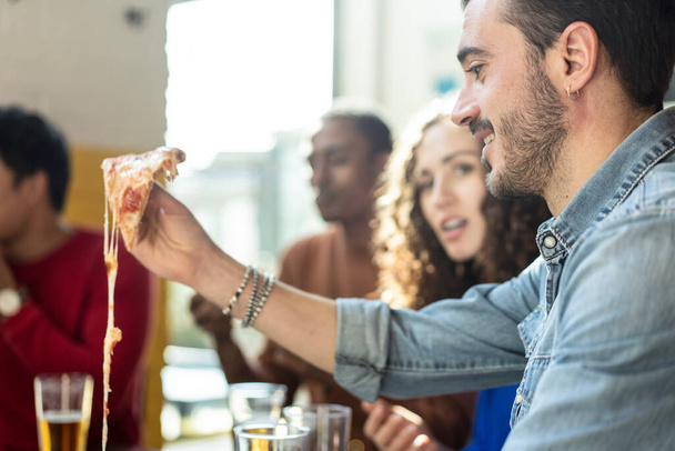 Smiling friends eating pizza at modern pizzeria restaurant - Friendship concept with multi ethnic people enjoying time together having fun at pizzeria with pizza and beer pints - Photo, Image