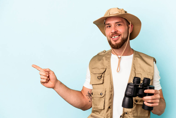 Young caucasian man looking at animals through binoculars isolated on blue background smiling and pointing aside, showing something at blank space. - Foto, afbeelding