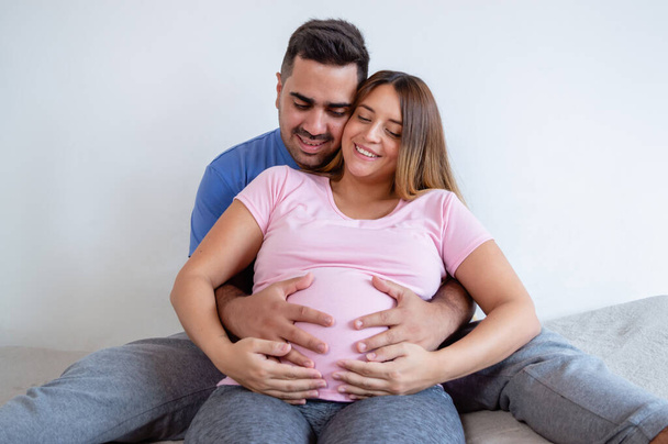 щаслива біла вагітна жінка з чоловіком сидить на ліжку посміхаючись і торкаючись дитини на животику, роблячи терапію вагітності і фізичні вправи
. - Фото, зображення