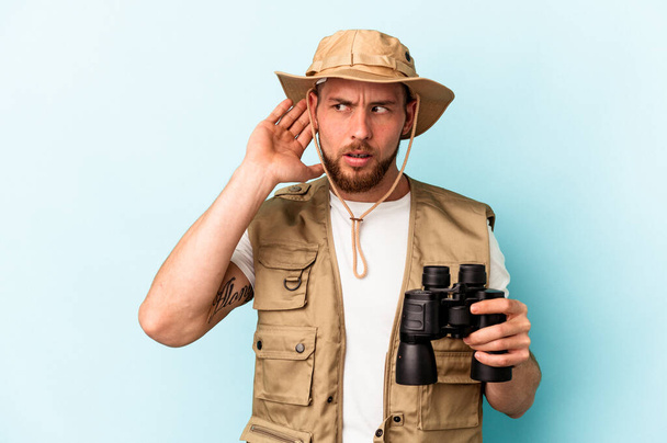 Young caucasian man looking at animals through binoculars isolated on blue background trying to listening a gossip. - Fotoğraf, Görsel