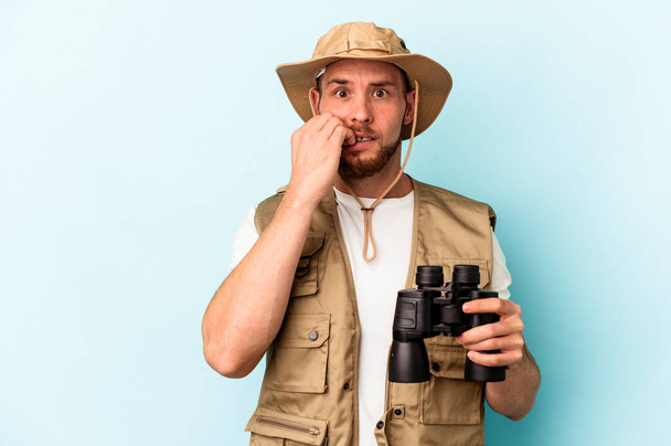 Young caucasian man looking at animals through binoculars isolated on blue background biting fingernails, nervous and very anxious. - Foto, imagen