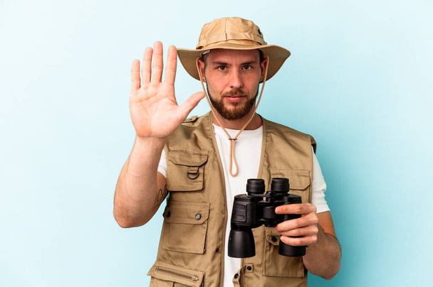 Young caucasian man looking at animals through binoculars isolated on blue background smiling cheerful showing number five with fingers. - Photo, image