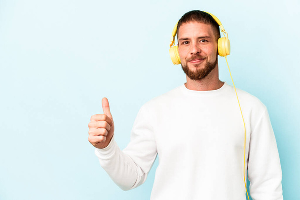 jonge Kaukasische man luisteren naar muziek geïsoleerd op blauwe achtergrond glimlachen en heffen duim omhoog - Foto, afbeelding