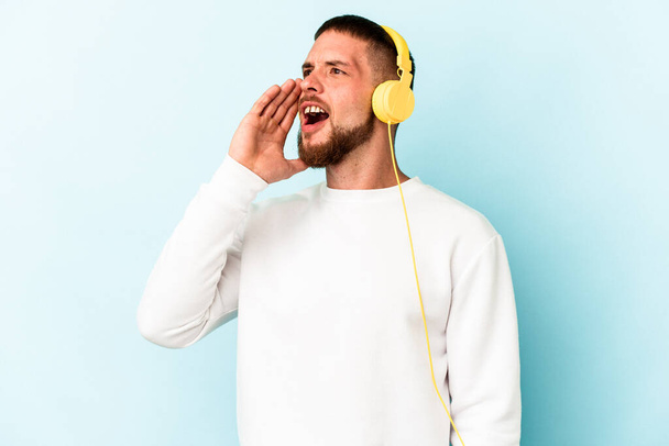 Junger kaukasischer Mann hört isoliert Musik auf blauem Hintergrund und hält Handfläche in der Nähe geöffneten Mundes. - Foto, Bild