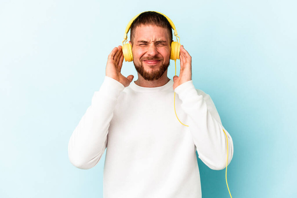 Joven hombre caucásico escuchando música aislada sobre fondo azul cubriendo oídos con las manos. - Foto, imagen