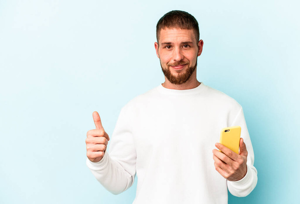 Молодой кавказский мужчина с мобильным телефоном на синем фоне улыбается и поднимает большой палец вверх - Фото, изображение