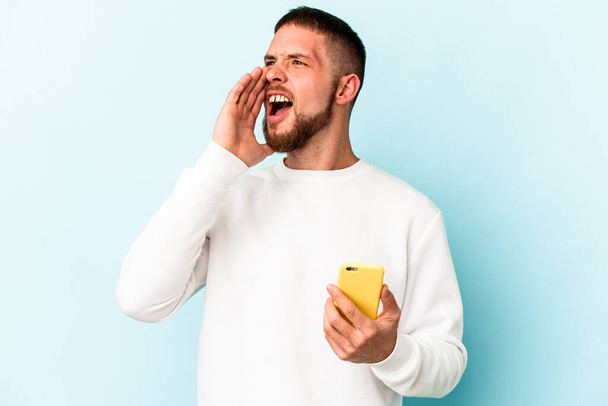 Junger kaukasischer Mann mit Handy isoliert auf blauem Hintergrund schreit und hält Handfläche in der Nähe geöffneten Mundes. - Foto, Bild