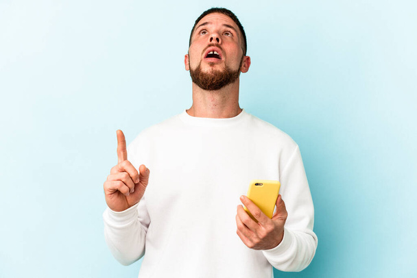 Junger kaukasischer Mann hält Handy isoliert auf blauem Hintergrund und zeigt kopfüber mit geöffnetem Mund. - Foto, Bild