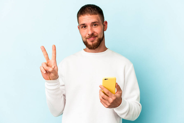 Junger kaukasischer Mann hält Handy isoliert auf blauem Hintergrund und zeigt Nummer zwei mit Fingern. - Foto, Bild