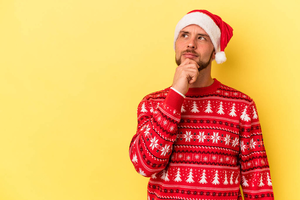 Young caucasian man celebrating Christmas isolated on yellow background looking sideways with doubtful and skeptical expression. - Fotoğraf, Görsel