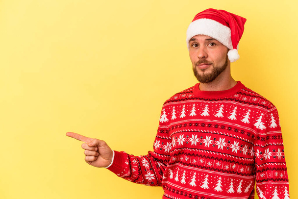 Junger kaukasischer Mann, der Weihnachten isoliert auf gelbem Hintergrund feiert, lächelnd zur Seite zeigt und etwas im leeren Raum zeigt. - Foto, Bild