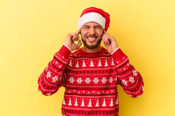 młody biały człowiek świętuje Boże Narodzenie odizolowany na żółtym tle przykrywając uszy z rąk. - Zdjęcie, obraz