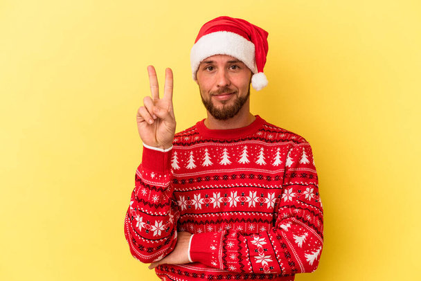 Junger kaukasischer Mann feiert Weihnachten isoliert auf gelbem Hintergrund und zeigt Nummer zwei mit Fingern. - Foto, Bild