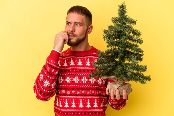 Junger kaukasischer Mann kauft Weihnachtsbäumchen isoliert auf gelbem Hintergrund und denkt entspannt über etwas nach, das einen Kopierraum betrachtet. - Foto, Bild