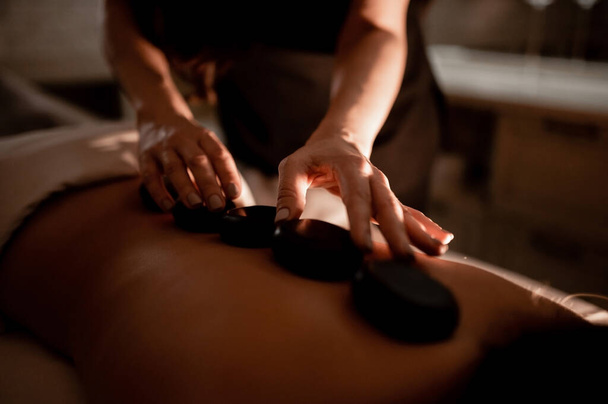 La masajista masajea la espalda de la chica con piedras calientes. Masaje de piedra caliente en el spa. - Foto, imagen