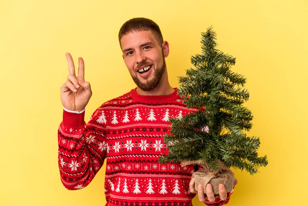 Junger kaukasischer Mann kauft Weihnachtsbäumchen isoliert auf gelbem Hintergrund und zeigt fröhlich und unbeschwert ein Friedenssymbol mit Fingern. - Foto, Bild