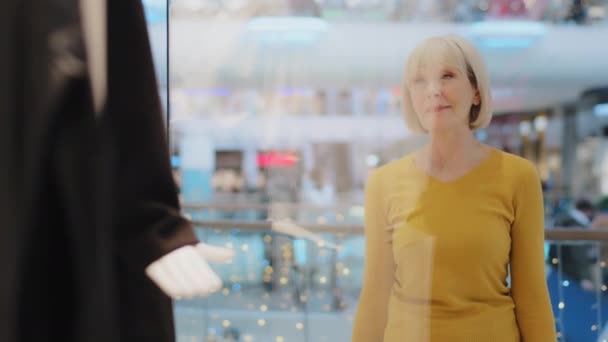 Mature middle aged caucasian woman looking sadly at showcase window clothing store in shopping mall upset frustrated sad because higher prices costly purchase luxury too expensive not affordable - Footage, Video