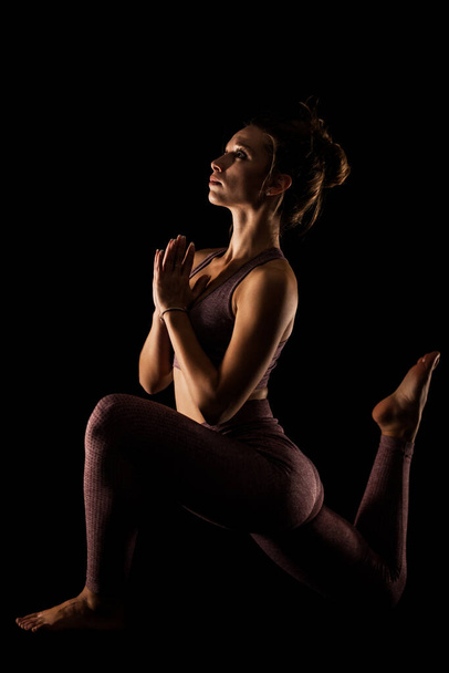 Mulher apta a praticar poses de ioga. Lado iluminado meia silhueta menina fazendo exercício em estúdio contra fundo preto. - Foto, Imagem