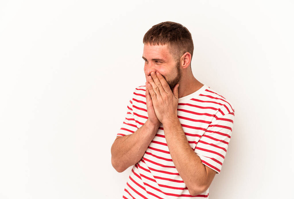 Молодой кавказский мужчина с диастемой на белом фоне смеется над чем-то, закрывая рот руками. - Фото, изображение