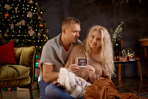 Boldog boldog terhes pár ölelkezik együtt ultrahanggal a babájukról. Esztétikai hangulatú otthon belső közelében karácsonyfa ünnepi dekorációk. Őszinte, igazi pillanat. Karácsonyi család - Fotó, kép