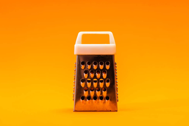Malý lesklý kovový drtič izolovaný na oranžovém pozadí, detailní záběr na sýr a nerez struhadlo. - Fotografie, Obrázek